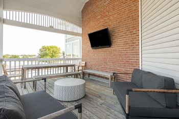 A balcony with a grey couch, a white ottoman and a flat screen tv mounted on a brick wall